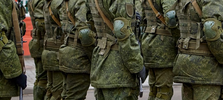 A group of soldiers dressed in green military uniforms march together in formation on a parade ground during a military ceremony. They are wearing black gloves and their faces are not visible.