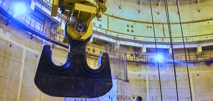 The image shows a large industrial crane hook hanging inside a vast, unfinished concrete structure. The hook appears to be lowered from the top of the building, suggesting heavy construction or lifting operations are in progress. The concrete walls are unfinished and the lighting in the structure is sparse, creating a sense of industrial scale and activity.