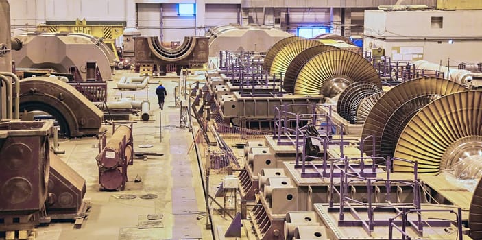 A lone worker walks through a vast factory floor, dwarfed by the imposing presence of massive turbine blades in various stages of assembly. The scene captures the raw power and intricate engineering involved in constructing these essential components of modern energy infrastructure.