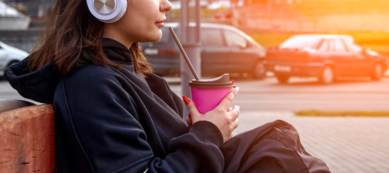 A young woman sits on a bench in an urban setting, enjoying a refreshing beverage on a warm afternoon. She wears headphones and seems engrossed in her own thoughts as she takes a break amidst the citys bustling energy. The sun shines brightly in the background, creating a warm and inviting atmosphere.