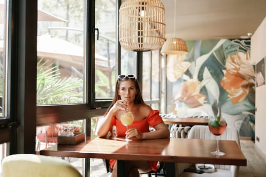 woman in a cafe seated, holding beverage. Dressed in an orange dress with her hair down. Bright interior, large windows allow natural light. Beverage consumption for refreshment