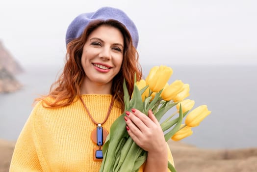 A woman in a yellow sweater holding a bouquet of yellow tulips. She is wearing a blue hat and a necklace