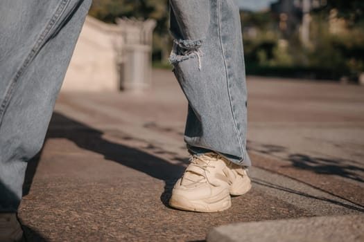 A woman is walking down a sidewalk wearing ripped jeans and a gray jacket. The image conveys a casual and relaxed atmosphere, as the woman is enjoying her walk