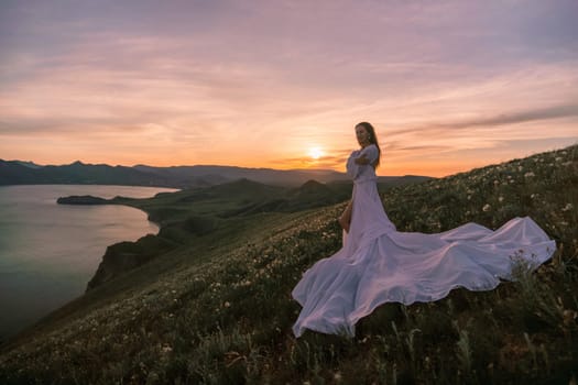 A woman in a long white dress stands on a hill overlooking a lake. The sky is a mix of orange and pink hues, creating a serene and romantic atmosphere. The woman's dress billows in the wind
