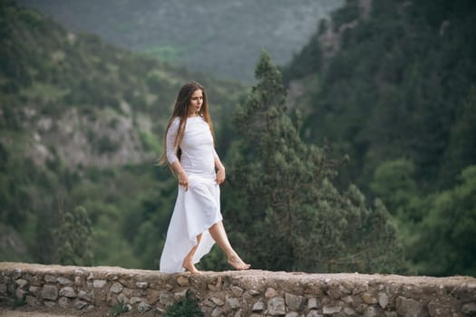 A woman in a white dress is walking on a stone wall. The image has a serene and peaceful mood, as the woman is alone and surrounded by nature. The dress and the setting create a sense of elegance