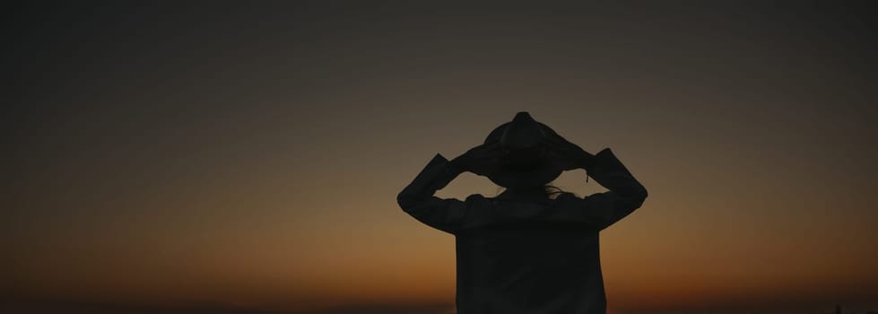 A woman is standing in the dark with her arms outstretched, looking up at the sky. The sky is a deep blue color, and the sun is setting. The woman is lost in thought