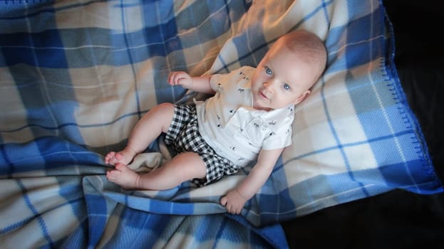 A baby in light clothes is lying on the bed. He is lying in a blue plaid blanket on a gray bed. High quality photo