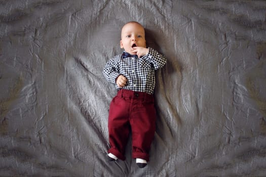 A baby in a plaid shirt and maroon pants lies on the bed. High quality photo