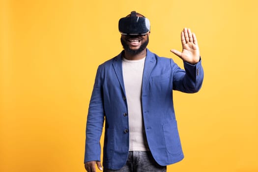 Smiling man wearing virtual reality headset, interacting with interface. Upbeat BIPOC person using high tech futuristic modern VR gadget, isolated over yellow studio background
