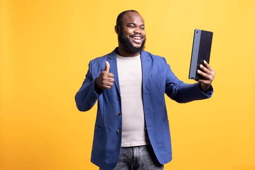 Cheerful man agreeing with friends during teleconference meeting using tablet, showing thumbs up sign, studio background. Joyous person doing positive hand gesturing with mates during videocall