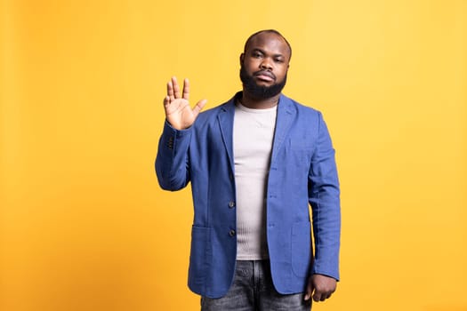 African american man reluctantly doing salutation hand gesture. Portrait of emotionless BIPOC person raising arm to greet someone, gesturing, isolated over yellow studio background