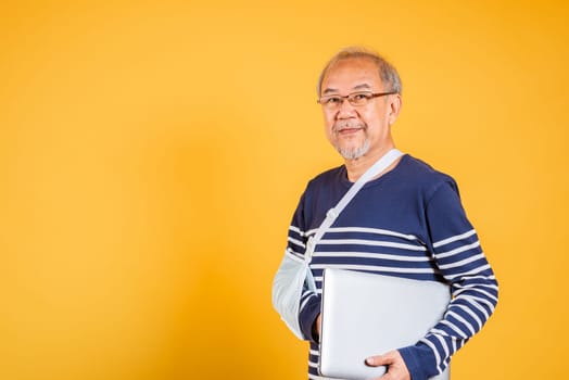 Portrait Asian old business man cast his arm hold laptop computer studio isolated yellow background, elderly man accident broken arm wear splint treatment while working, dedication and recovery