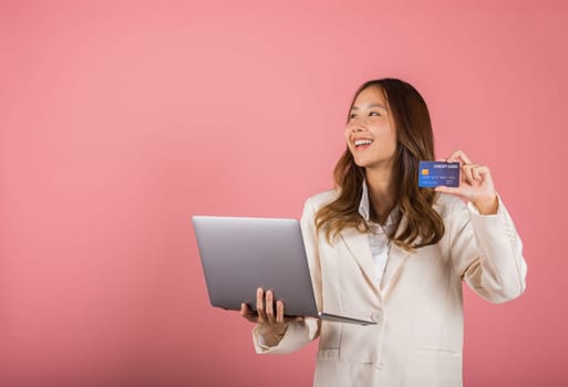 Portrait of Asian beautiful young woman customer showing credit card for online payment shopping transactions cashback loan on laptop studio shot isolated on pink background