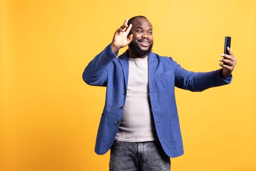 Cheerful man using smartphone to take selfies and post them on social media. Happy BIPOC person taking photos using phone selfie camera, isolated over yellow studio background