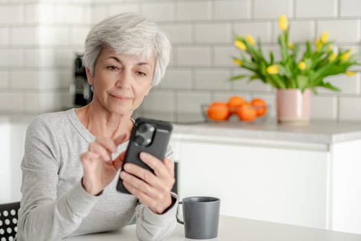 Grey-Haired Attractive Woman Searches For Information On Phone At Home At Table