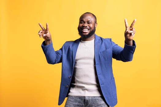 Portrait of cheerful man doing victory sign after winning, isolated over studio background. Lively radiant BIPOC person celebrating achievement, doing victorious hand gesturing