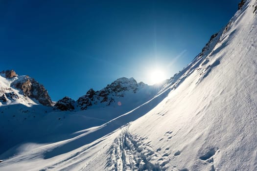 Landscape of a frozen mountainside with the sun setting behind the rocky peaks - a mountaineering trail winding through snowy terrain.