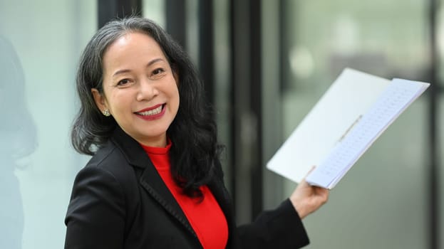 Attractive mature businesswoman holding documents and smiling confidently at camera.