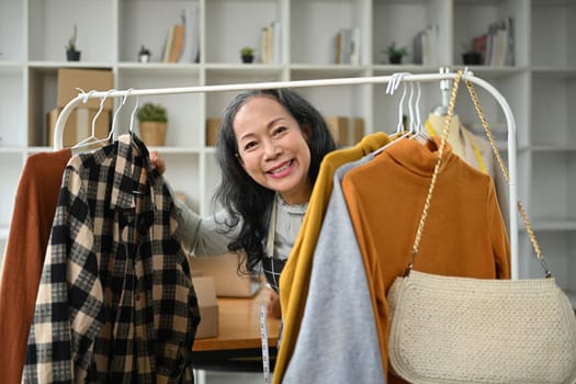 Middle age asian woman small business owner standing near clothing rack and smiling cheerfully.