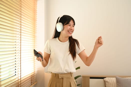 Cheerful young asian woman dancing while enjoying music through headphones in living room.