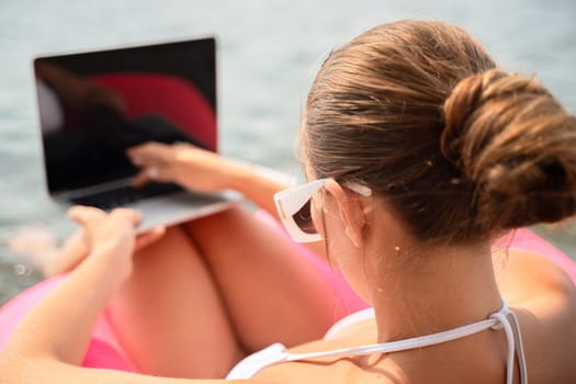 A woman is sitting in a pink inflatable raft with a laptop on her lap. She is wearing sunglasses and she is enjoying her time by the water