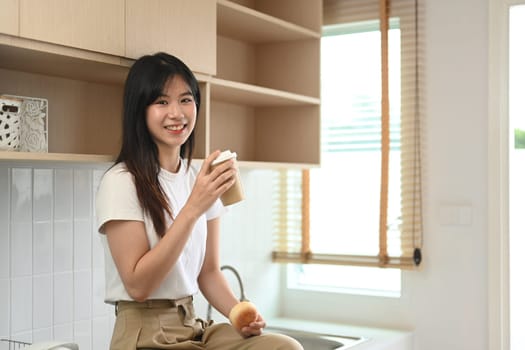 Portrait of cheerful young Asian woman having breakfast in minimal kitchen interior.