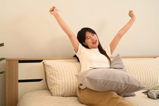 Carefree young Asian woman wearing casual clothes stretching her arms in bed.