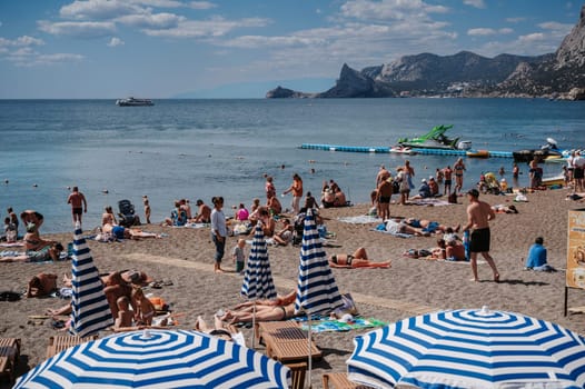 SUDAK, CRIMEA - SEPTEMBER 6, 2021: People sunbathe on beach on Black Sea coast on beach in resort town of Sudak in Crimea