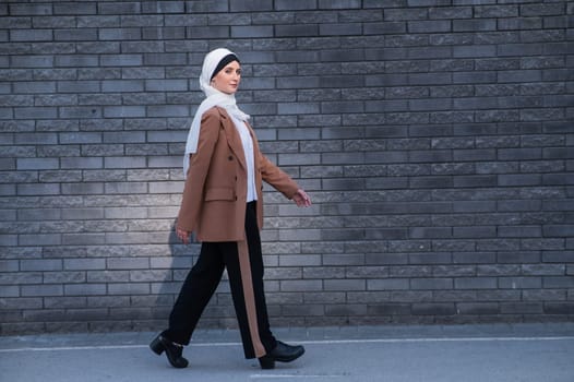 A young woman dressed in a hijab and a business suit walks along a brick wall