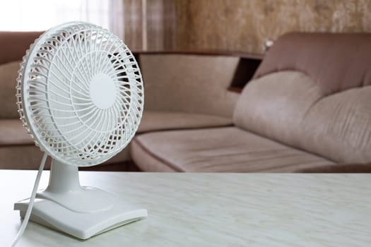 Tabletop compact white plastic fan with included blades disperses cold air throughout the home room against the backdrop of the sofa on hot day