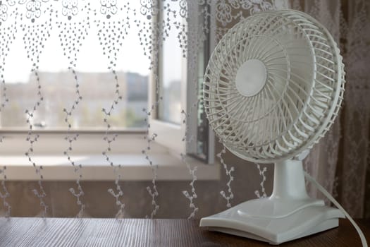 White table fan with rapidly rotating blades to cool the air in home room against background of window on hot sunny day, increasing the temperature in room using electric fan