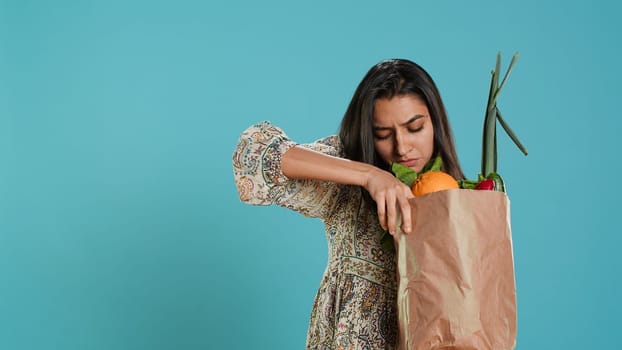 Indian woman with ecological paper bag in hands looking at bell pepper, living healthy lifestyle. Ecology lover holding shopping bag with organic produce, studio backdrop, camera A