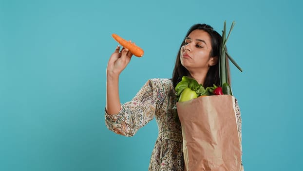 Woman with paper bag in hands filled with vegetables checking carrot for impurities. Conscious living customer with purchased groceries to be used as cooking ingredients, studio background, camera A