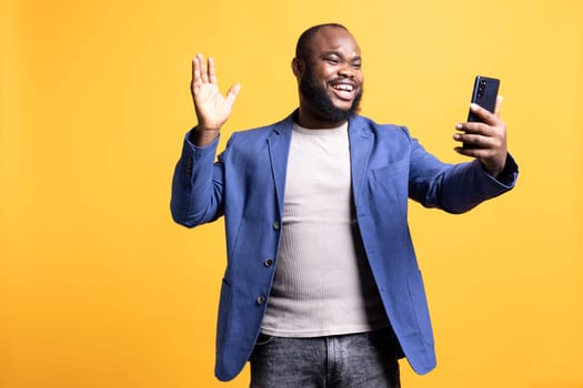 Cheerful man excited to greet friends during teleconference meeting using smartphone, studio background. Smiling person waving hand, saluting mate during internet video call on mobile phone