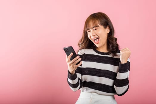 Happy Asian portrait beautiful cute young woman teen smiling excited using mobile phone say yes Studio shot isolated on pink background, Thai female surprised make winner gesture on smartphone