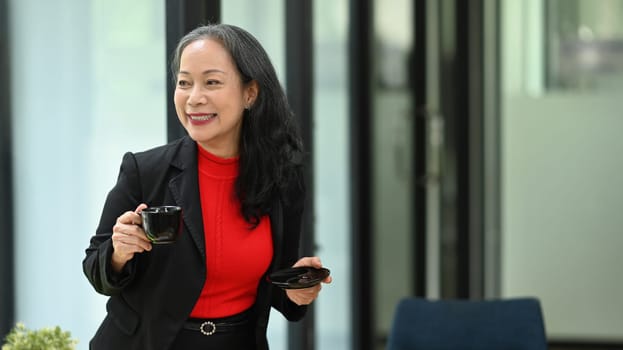 Charming mature businesswoman taking break from work, enjoying hot drink in her office.