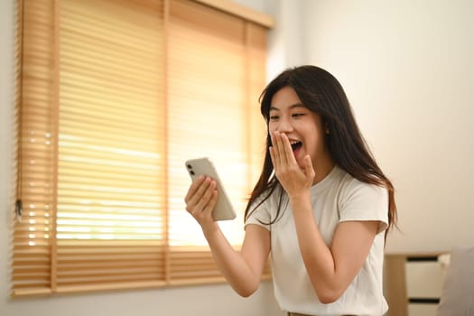 Shocked young asian lady looking at smartphone screen excited by unexpected notification.