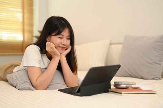 Cute young asian woman lying in comfortable bed and using digital tablet.