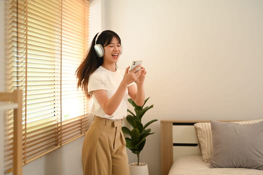 Joyful young woman holding phone listening to favorite songs and dancing in living room.