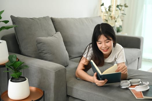 Pretty young Asian woman reading book while lying on couch at home.