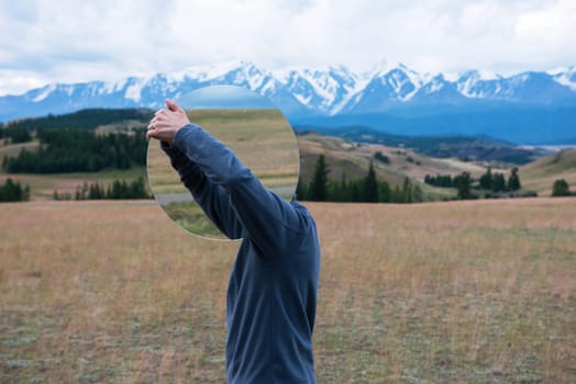 Man standing in in summer Altai mountains in Kurai steppe and holding circle mirror. Creative travel concept