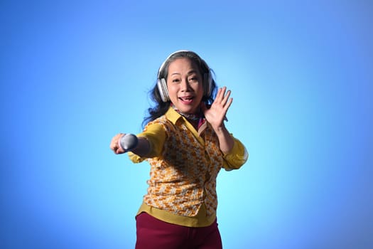 Energetic elderly woman in headphones singing with microphone over light blue background.