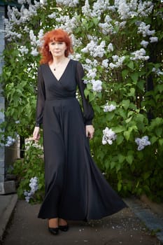 Elegant senior mature Woman in Black Dress by Blooming Lilac Bush at Dusk. Woman with red hair stands poised among lilac blooms