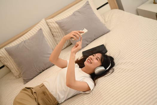 Smiling young woman listening to music with headphones and using smartphone while lying in bed.