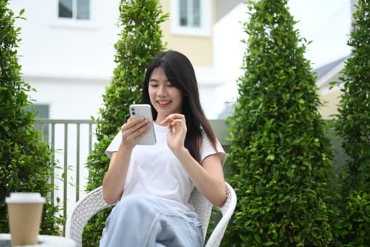 Happy young asian woman using mobile phone relaxing in the garden surrounded by plants.