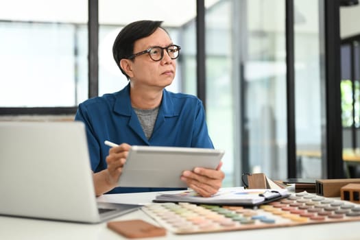 Thoughtful interior designer sitting at creative workplace with digital tablet and looking away.