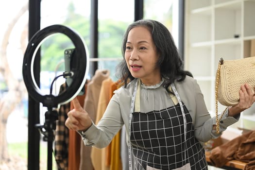 Smiling mature woman online seller showing clothes in front of smartphone while live streaming.