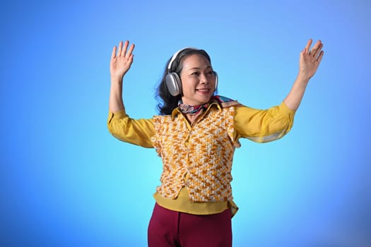 Cheerful senior woman in headphones dancing with hand raised over blue background.
