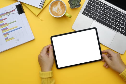 Top view woman holding digital tablet on yellow background with laptop and financial documents.