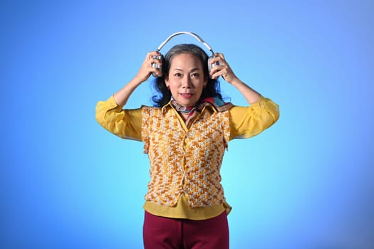 Portrait of beautiful elderly woman wearing headphone standing against light blue background.